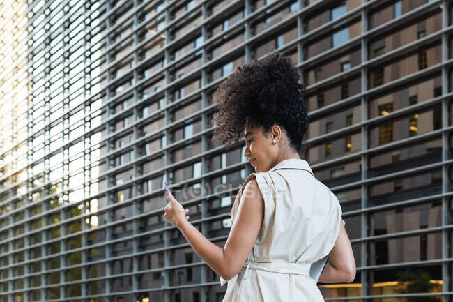 Вид на молодую афроамериканку с вьющимися волосами, смотрящую на мобильный телефон, пронося нетбук мимо современного здания — стоковое фото