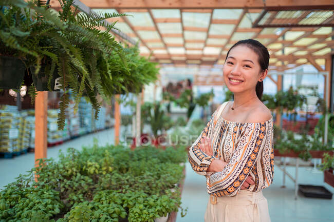 Удовлетворенная молодая азиатка-покупательница со сложенными руками смотрит в камеру на горшечные растения в садовом магазине — стоковое фото