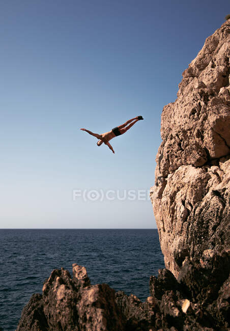Active male athlete in swimming trunks jumping from mount into sea under light sky in Ibiza Spain — Stock Photo