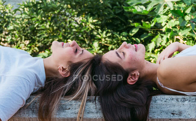 Side view of smiling female teen with acne lying against best friend with closed eyes on bench in park — Stock Photo