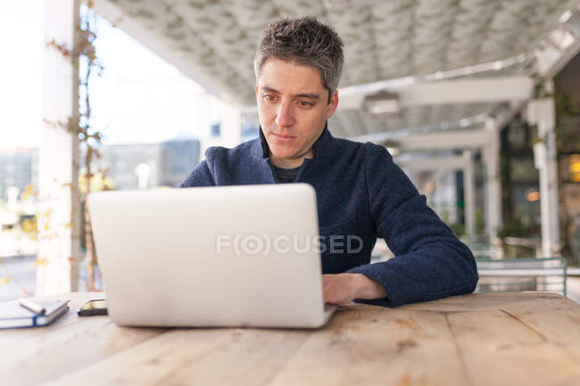 Jeune homme en tenue tendance parcourant le netbook tout en travaillant sur un projet dans la rue — Photo de stock