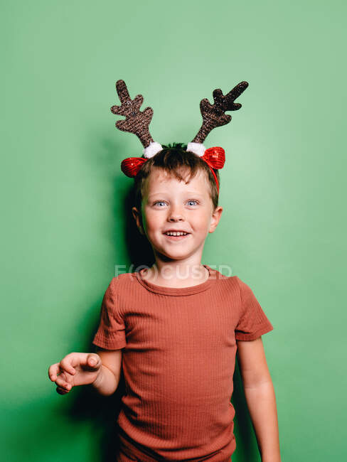 Sorrindo menino vestindo chifres de rena headband de pé contra fundo verde e olhando para a câmera — Fotografia de Stock