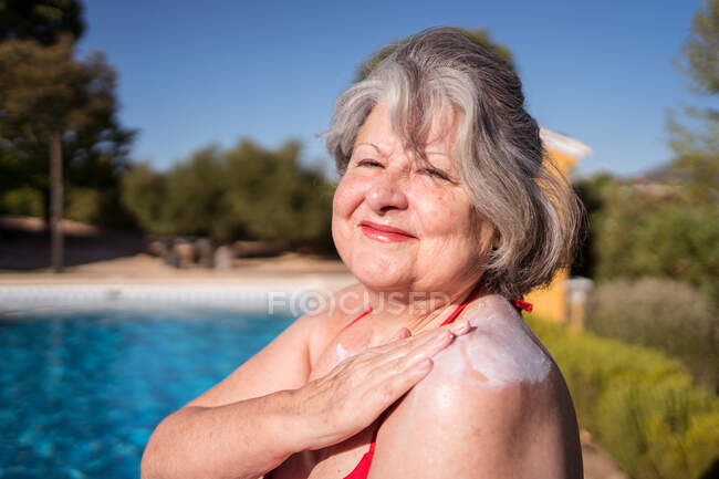 Vista laterale della donna spensierata con capelli grigi che applica la protezione solare sulla spalla mentre si gode la giornata di sole a bordo piscina — Foto stock
