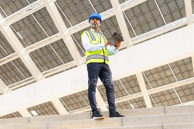 Нижній кут середнього віку інженер-латиноамериканець у формі з планшетом, що стоїть далеко під сонячною електростанцією. — стокове фото