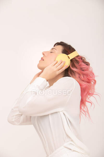 Vista lateral de mujer tranquila con pelo rosa en blusa blanca de pie con los ojos cerrados y escuchando música en auriculares sobre fondo claro - foto de stock