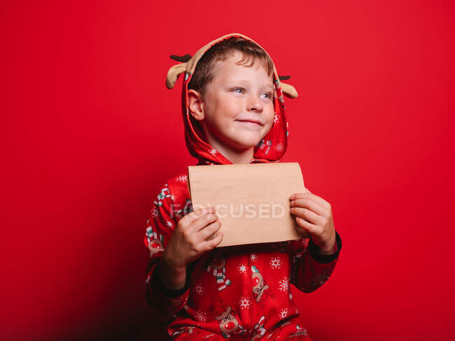 Menino feliz traje festivo olhando para longe ao ler carta contra fundo vermelho durante a festa de Natal — Fotografia de Stock