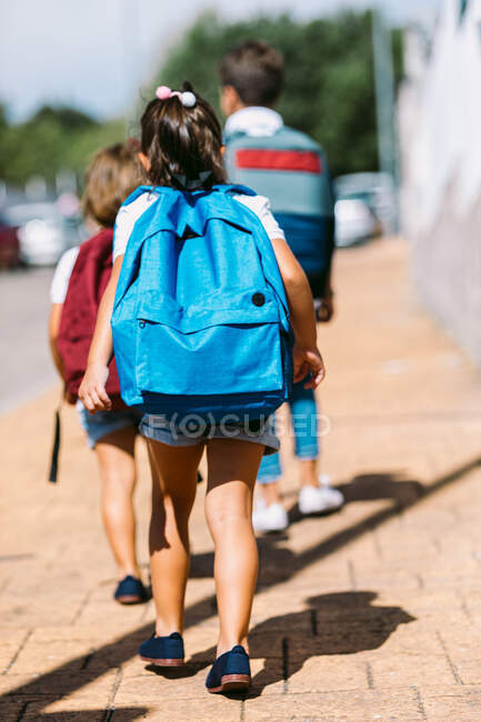 Rückansicht gesichtsloser Schulkinder mit Rucksäcken, die an sonnigen Tagen auf gefliestem Bürgersteig in der Stadt spazieren — Stockfoto