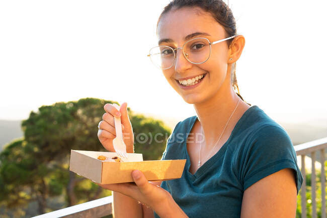 Vista lateral de la joven hembra feliz mirando a la cámara mientras come sabrosos gofres belgas con crema batida en caja de comida para llevar contra monturas en la espalda iluminada - foto de stock