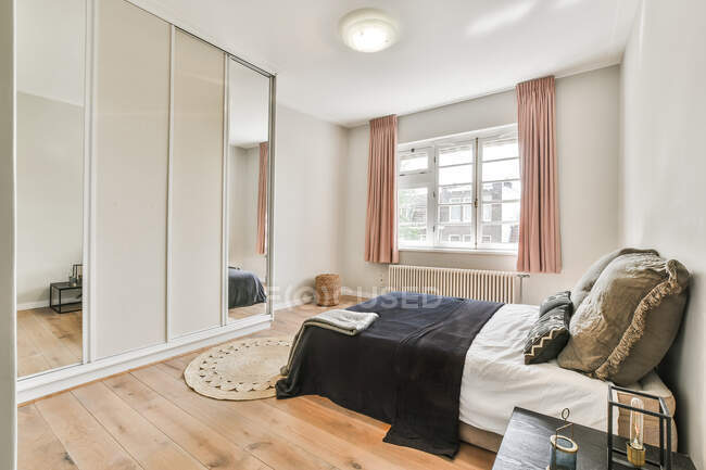 Zeitgenössisches helles Schlafzimmer mit bequemem Bett mit Kissen und Kleiderschrank mit verspiegelter Oberfläche in Fensternähe — Stockfoto