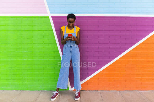 Full body of African American female in denim overalls standing near colorful wall and browsing cellphone — Stock Photo