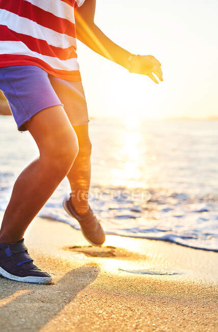 Colheita menino irreconhecível correndo de ondas de lavagem molhado costa arenosa iluminada pela luz solar brilhante ao pôr do sol — Fotografia de Stock