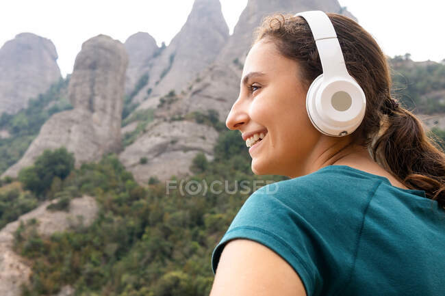 Joven turista soñadora mirando hacia otro lado disfrutando de la canción desde auriculares inalámbricos contra Montserrat y árboles en España - foto de stock