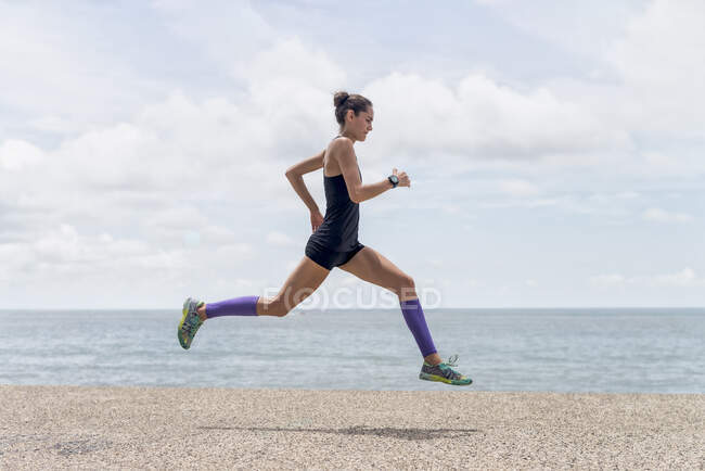 Vista lateral do corredor fêmea apto no momento de saltar sobre o calçadão durante o treinamento cardio energético no verão — Fotografia de Stock