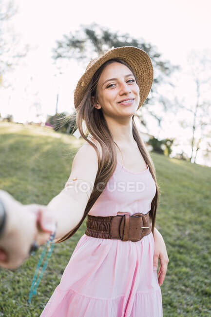Adolescente souriante en robe de soleil et chapeau de paille tenant la récolte partenaire anonyme à la main tout en regardant la caméra dans le parc — Photo de stock