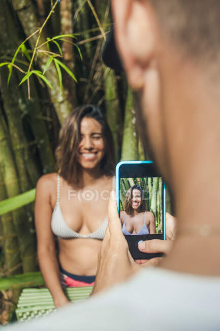 Recorte viajero masculino anónimo tomando la foto de la hembra sonriente amada en el teléfono celular contra las plantas de bambú en la luz del día - foto de stock