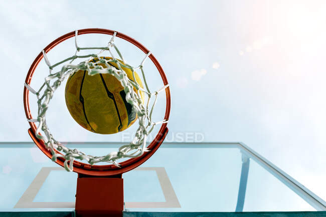 Von unten fliegt der Ball in den Basketballkorb, der während des Spiels am Backboard vor blauem Himmel auf dem öffentlichen Sportplatz befestigt ist — Stockfoto