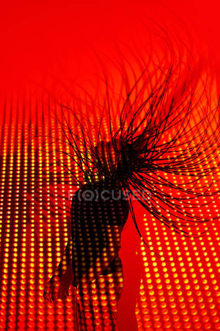 Vue latérale de femmes millénaires ethniques méconnaissables secouant de longs cheveux tressés tout en dansant près du mur avec un éclairage au néon rouge vif — Photo de stock