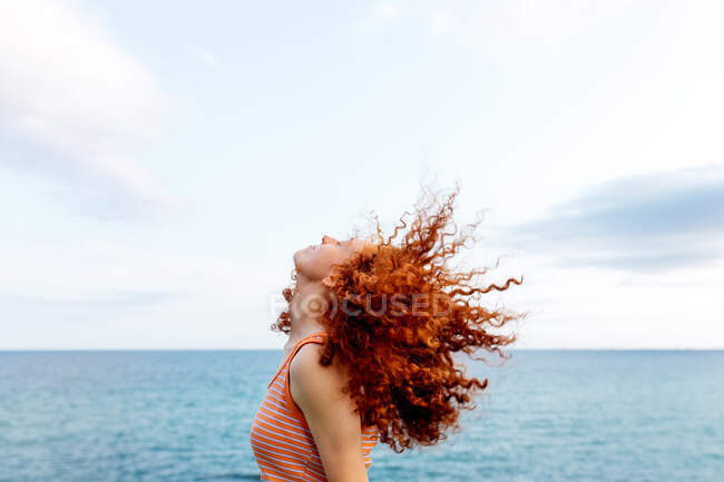 Side view of carefree anonymous female with eyes closed shaking curly ginger hair on coast of blue sea — Stock Photo