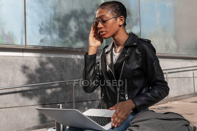 Conteúdo Freelancer afro-americano em jaqueta de couro sentado com netbook em voltas durante o trabalho on-line na rua, enquanto ele coloca seus óculos — Fotografia de Stock
