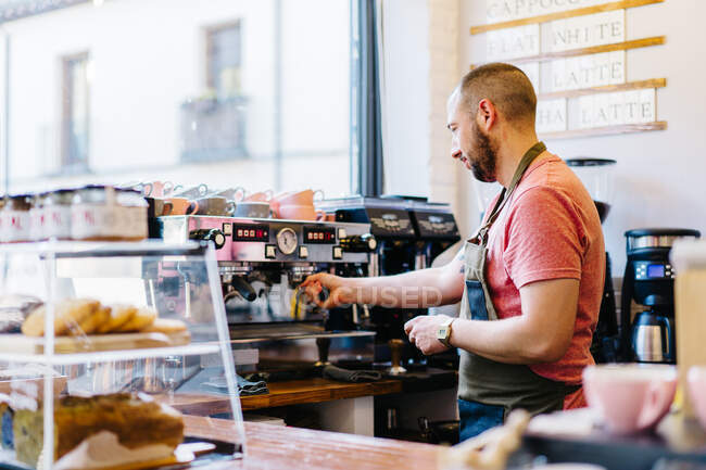 Side view of male barista in apron with portafilter brewing coffee using coffee machine while standing near counter in coffee house — Stock Photo