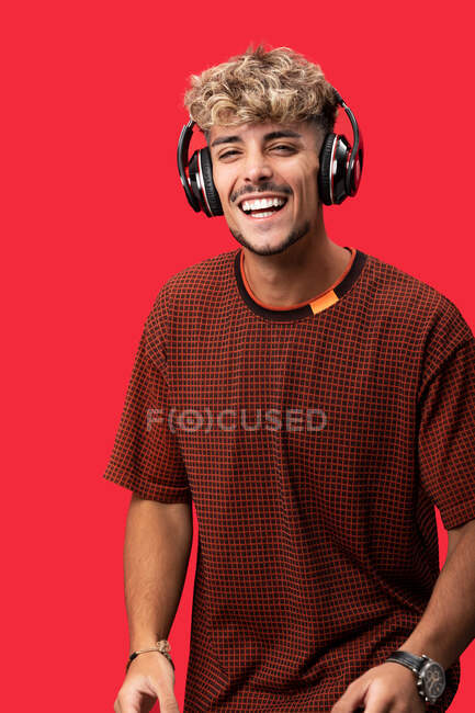 Positiver junger Mann mit lockigem Haar, der breit lächelt, während er die Playlist in drahtlosen Kopfhörern vor rotem Hintergrund genießt — Stockfoto