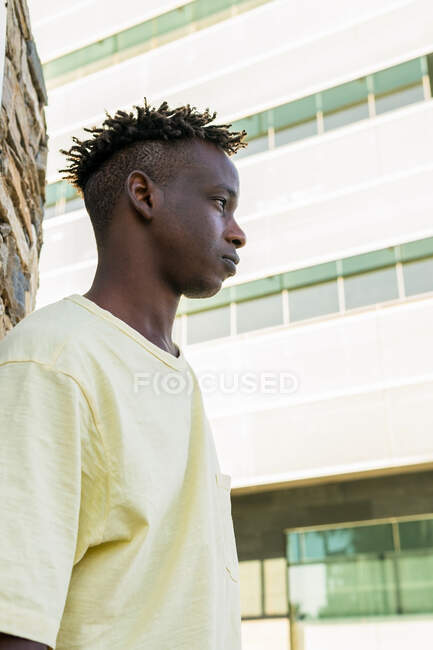 D'en bas jeune homme afro-américain sans émotion portant des vêtements décontractés debout près du mur de pierre — Photo de stock