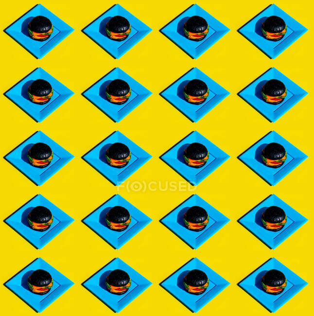 Зверху чорні гамбургери на квадратних синіх пластинах утворюють безшовний візерунок на жовтому фоні — стокове фото
