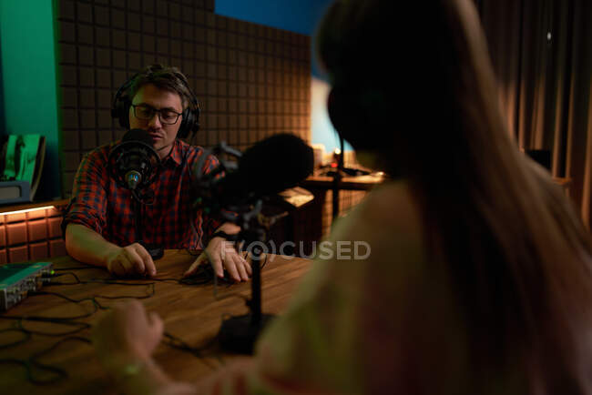 Jeune animateur radio masculin en chemise à carreaux et écouteurs assis à table avec microphone et communiquant avec un collègue anonyme lors de l'enregistrement de podcast en studio — Photo de stock