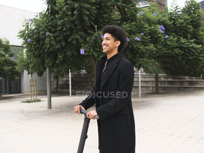 Trendiger junger Afroamerikaner mit dunklem lockigem Haar in stylischem Outfit steht auf dem Stadtplatz und schaut nach dem Rollerfahren weg — Stockfoto