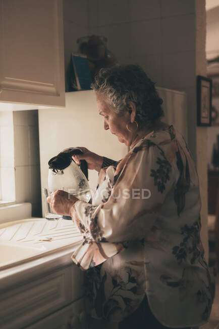 Side view of concentrated elderly woman pouring hot water from transparent electric kettle into glass tea mug while making drink with teabag in kitchen — Stock Photo