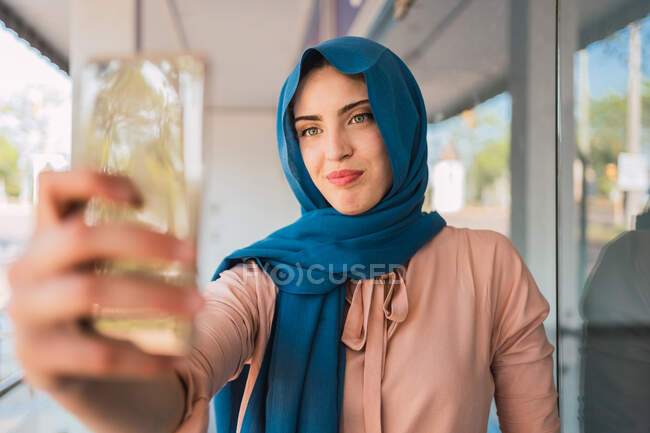 Alegre mujer árabe en el hiyab tradicional de pie en la calle de la ciudad y tomando auto disparo en el teléfono móvil - foto de stock