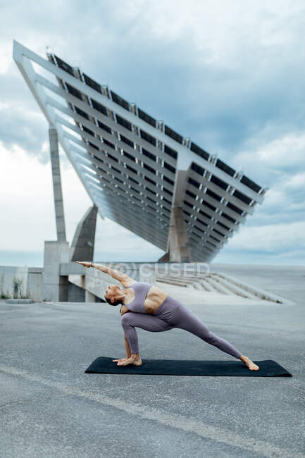Cuerpo completo de mujer descalza deportiva haciendo postura Utthita Parshvakonasana mientras practica yoga en la calle cerca del panel solar en la ciudad - foto de stock