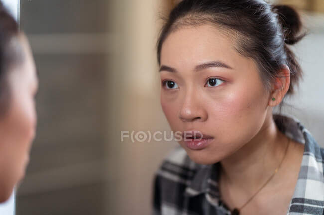 Attraktive junge Asiatin in lässigem Hemd blickt emotionslos in Spiegel im hellen Raum auf verschwommenem Hintergrund — Stockfoto