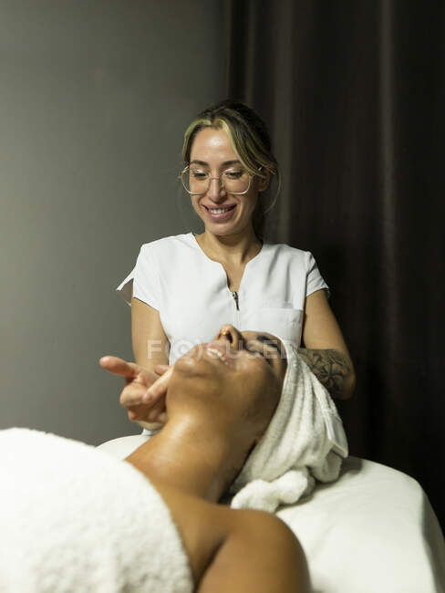Masseur féminin utilisant des masseurs tout en massant le visage du client en serviette sur le canapé dans un centre de spa lumineux — Photo de stock