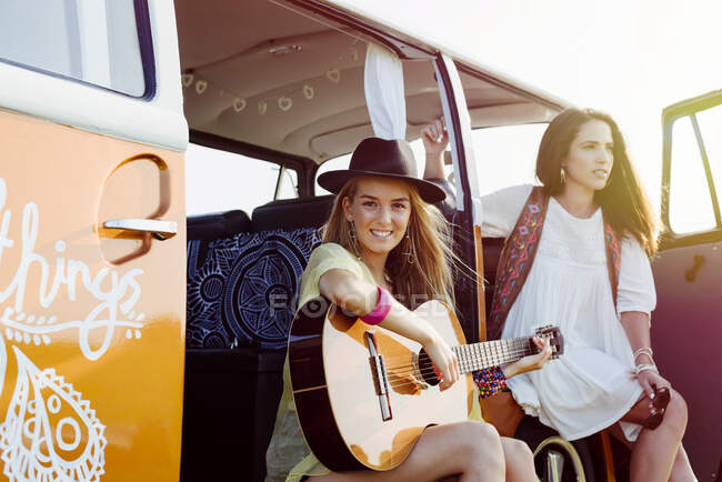 Две симпатичные девушки, одетые в летнюю одежду, веселятся у фургона и играют на гитаре — стоковое фото