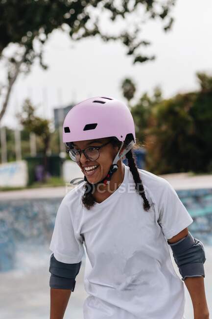 Улыбающаяся молодая афроамериканка в очках в светло-лиловом защитном шлеме и серых прокладках на коньках в парке — стоковое фото