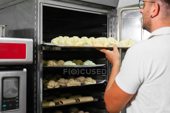 Young stylish male baker in casual clothes and eyeglasses putting croissant of raw dough on metal rack while baking bread in kitchen — Stock Photo