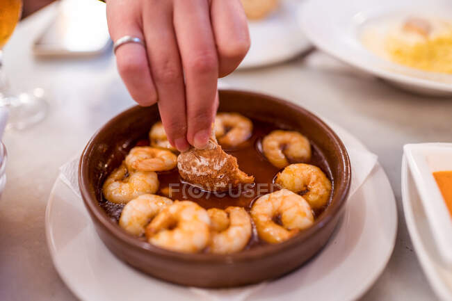 From above of crop unrecognizable woman dipping bread piece into sauce of appetizing traditional gambas al ajillo dish served in bowl in restaurant — Stock Photo