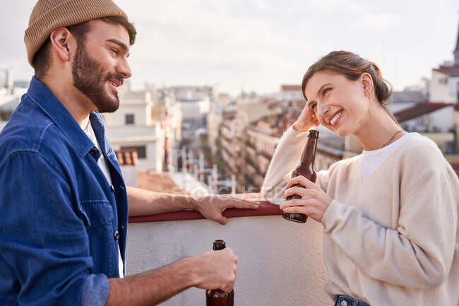 Full body of cheerful friends raising bottles of beer while laughing on balcony in old city — Stock Photo