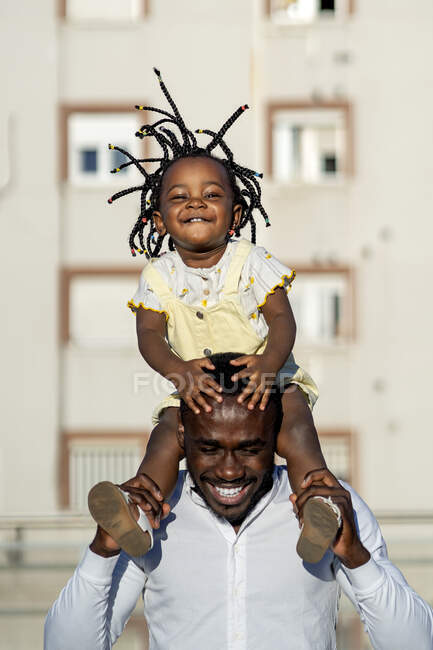 Fröhlicher afroamerikanischer Vater im Hemd, der seine kleine Tochter auf den Schultern trägt und springt, während er die Zeit zusammen auf der Straße in der Stadt im Sonnenlicht verbringt — Stockfoto