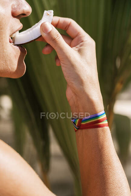 Vista lateral de la cosecha irreconocible persona con cinta de arco iris en la mano morder rebanada de coco natural fresco contra las plantas verdes en el tiempo de verano - foto de stock
