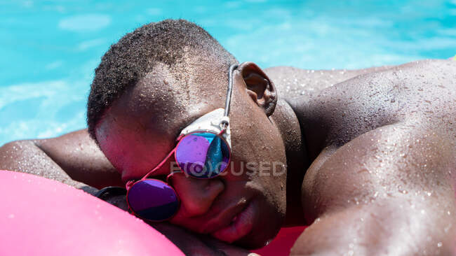 Uomo afroamericano senza camicia con occhiali da sole appoggiato su materasso gonfiabile rosa in piscina mentre prendeva il sole nella soleggiata giornata estiva — Foto stock