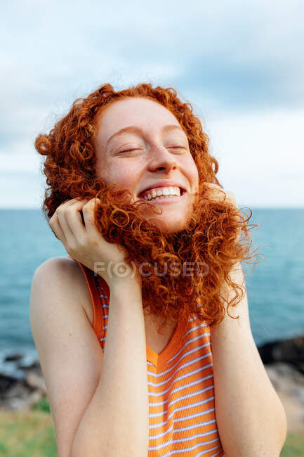 Joven pelirroja alegre haciendo pose infantil con el pelo rizado mientras disfruta de la libertad en la orilla del mar con los ojos cerrados - foto de stock