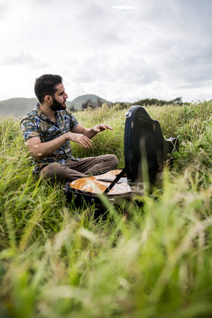 Вид збоку на спокійного чоловіка-музиканта в повсякденному одязі, що сидить на зеленій траві і відкриває чорний випадок акустичної гітари на березі моря в денний час — стокове фото