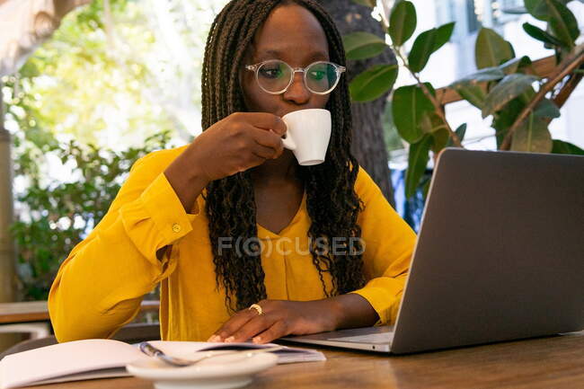 African American person drinking hot coffee sitting at wooden table in modern cafeteria while working in laptop on blurred background — Stock Photo