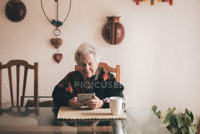 Mujer anciana sonriente usando ropa de abrigo sentada en la mesa con la tableta y la taza de té mirando a la pantalla - foto de stock