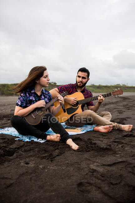 Full length of smiling man playing acoustic guitar with positive female friend playing ukulele while sitting on sandy coast in nature in cloudy day — Stock Photo
