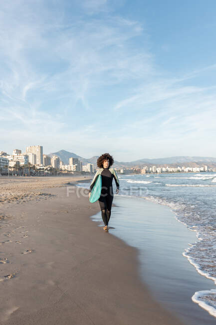 Joven surfista femenina reflexiva en traje de neopreno con tabla de surf caminando mirando a la orilla del mar bañada por el mar ondulante - foto de stock