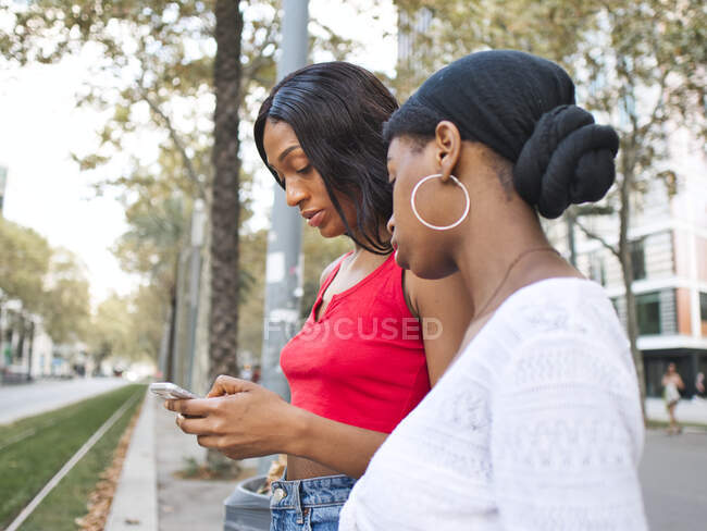 Vista lateral da concentrada afro-americana amigos do sexo feminino surf internet no celular, enquanto de pé na rua perto da estrada com a construção — Fotografia de Stock