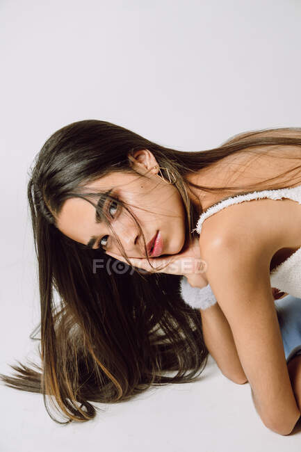 Vista lateral de la joven hispana con el pelo largo tocando la cara mientras se inclina hacia adelante y mirando a la cámara en el suelo - foto de stock